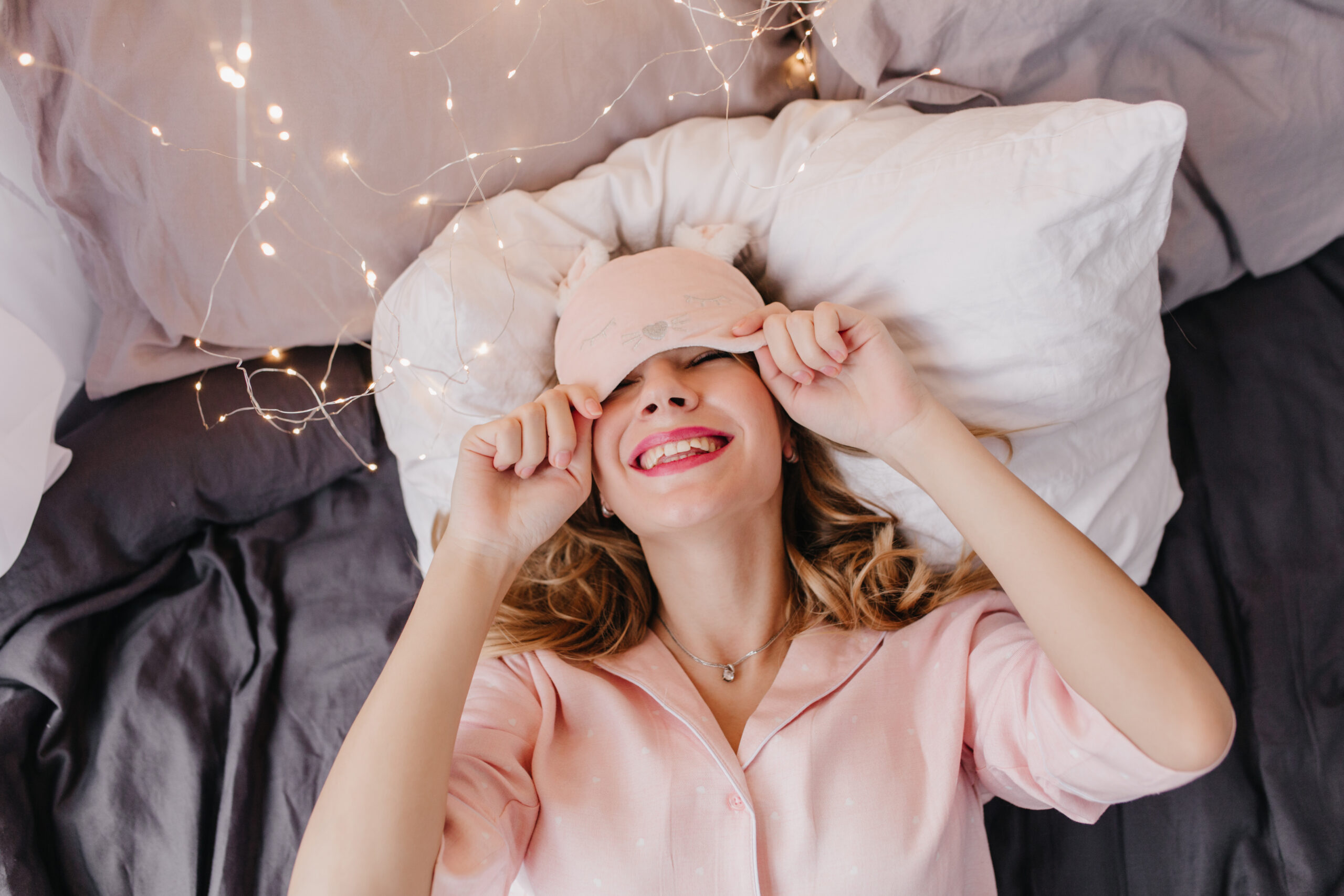 Lovable young woman in pink night-suit posing in bed with smile. Excited caucasian girl in eyemask relaxing in morning and laughing.